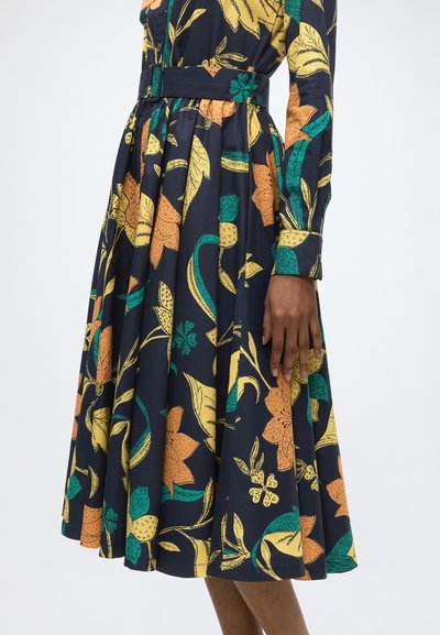 Femme portant une robe bleu marine avec un motif floral jaune, orange et vert et une ceinture en tissu assortie à la taille.