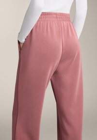 Růžové tepláky s volným střihem, elastickým pasem, bočními kapsami a hladkou strukturou. Tkanina vypadá měkce a mírně leskle.