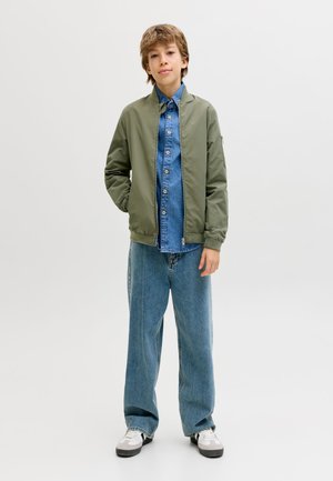 Poiss seisab käed osaliselt taskutes, seljas oliivroheline bomber-tagi sinise teksariietuse särgi ja laiade siniste teksapükstega, valgete tossudega.