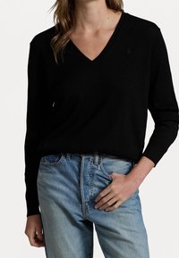 Svart v-ringad stickad tröja med långa ärmar, kombinerad med ljusblå raka jeans. Tröjan har en subtil logotyp på bröstet.