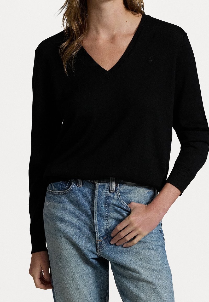 Svart v-ringad stickad tröja med långa ärmar, kombinerad med ljusblå raka jeans. Tröjan har en subtil logotyp på bröstet.