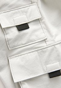 Primo piano di tessuto grigio chiaro con due tasche rettangolari, ciascuna fissata da una patta e da una linguetta intrecciata nera.