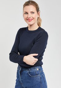 Pull en maille bleu marine à manches longues, avec un col rond et trois boutons à l'épaule. Associé à un jean bleu en denim taille haute.