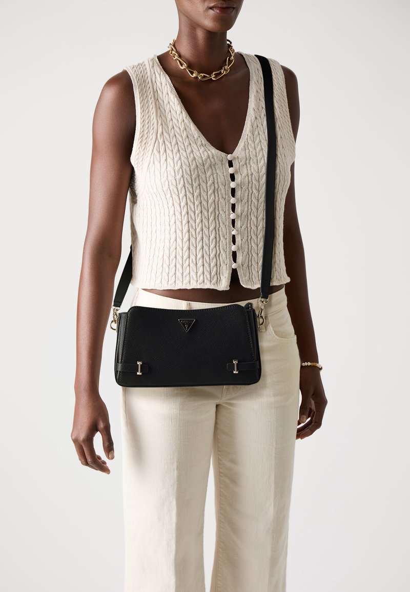 Zwarte textuur crossbody tas met gouden hardware en logo-details. Vergezeld van een crèmekleurig gebreid mouwloos topje en lichte broek.