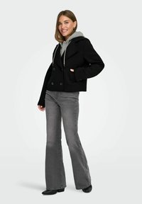 Žena se smiješi u crnom dvorednom kaputu preko sivoj dukserici, sivi razrezani traperice i crnim cipelama s špicastim vrhom, stoji na bijeloj pozadini.