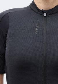 Κοντινή λήψη μαύρου υφασμάτινου ποδηλατικού τζέρσεϊ με κοντά μανίκια, φερμουάρ στο μπροστινό μέρος και διακριτικό κάθετο λογότυπο "Isadore" στο στήθος.