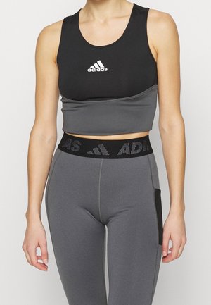 Femme portant un crop top Adidas noir et gris et un legging gris taille haute avec une ceinture Adidas noire, debout devant un fond uni.