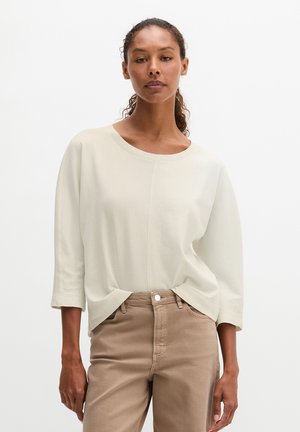 Vrouw met krullend haar draagt een losse witte top met lange mouwen en beige hoge taille broek tegen een effen witte achtergrond.