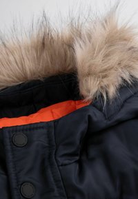 Czarna nylonowa kurtka z kołnierzem, który ma beżowe futro i pomarańczową podszewkę. Tkanina ma gładką fakturę z pikowanym wykończeniem.