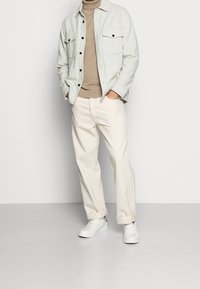 Camisa de botones gris claro con dos bolsillos en el pecho, jersey de cuello alto beige, pantalones blanco hueso y zapatillas blancas de textura lisa.