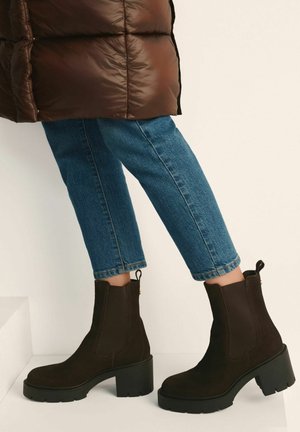 Brązowe zamszowe botki za kostkę z grubą czarną gumową podeszwą, elastycznymi bocznymi panelami i pętelką z tyłu, zestawione z niebieskimi krótkimi dżinsami.