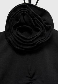 Haut en tissu noir avec un grand détail de fleur superposée au niveau du col, doté d'une texture lisse et de plis doux.