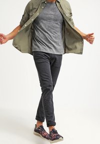 Outfit casual composto da una maglietta grigia sotto una camicia verde oliva aperta, pantaloni neri a sigaretta e scarpe scure con motivo floreale.