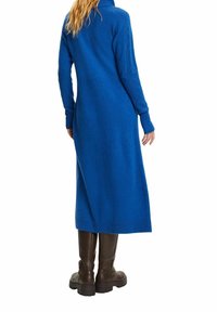 Femme portant une longue robe bleue en maille et des bottes brunes montant jusqu'aux genoux, debout de dos devant un fond blanc.