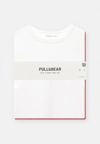Bijela majica s okruglim izrezom, preklopljena, s crvenim rubom na dnu, s bijelim etiketom na kojoj je ispisano ime brenda Pull&Bear.