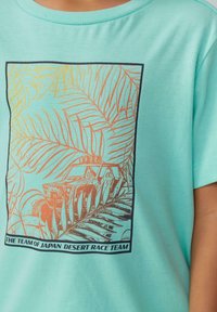 Türkises T-Shirt mit Rundhalsausschnitt, das ein gold- und orangefarbenes Wüstenmotiv zeigt, einschließlich Palmblättern und einer Fahrzeugillustration.