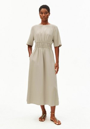 Femme portant une robe midi beige avec une taille élastique et des manches courtes, debout, mains dans les poches, associée à des sandales marron à lanières autour de la cheville.