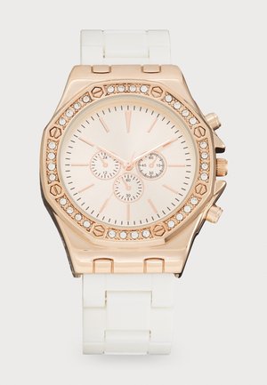 Montre analogique en or rose avec une lunette incrustée de cristaux, un cadran blanc présentant trois sous-cadrans, et un bracelet blanc à maillons.