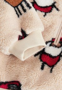 Mäkká béžová fleece tkanina s červeným a čiernym kresleným motívom, s ribovaným svetlobéžovým manžetovým lemom a hladkým detailom na zips.