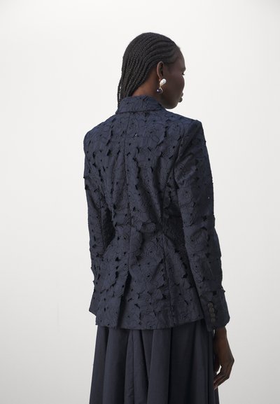 Navy blue embroidered blazer with floral lace pattern, tailored shape, and button accents, featuring textured fabric and a structured collar.