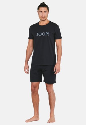 Mężczyzna stojący boso, ubrany w czarny t-shirt JOOP! i pasujące czarne spodenki na jednolitym białym tle.