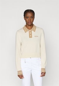 Γυναίκα με πλεξουδάκια, ντυμένη με κρεμ πουλόβερ με μπεζ γιακά και μανσέτες, και λευκό παντελόνι με μικρό αλυσίδα αξεσουάρ.