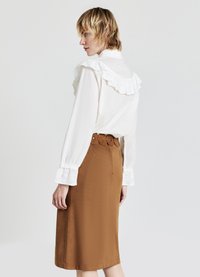 Camicia bianca con colletto a volant, maniche lunghe; gonna a matita marrone con dettaglio della cintura rotonda e accenti metallici circolari. Texture liscia complessivamente.