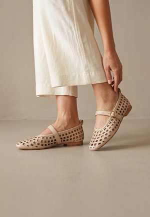 Persona que lleva pantalones cortos de color beige claro y zapatos planos Mary Jane tejidos en beige claro con una correa de hebilla, tocando un zapato.