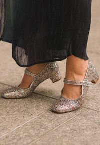 Zapatos Mary Jane de color plateado brillante con un tacón bajo, que presentan un corte en la puntera y una correa en el tobillo. La superficie tiene una textura de lentejuelas multicolores.