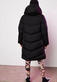 Czarna kurtka puchowa z wzorem w zygzak, oversize'owy kaptur i miękka tekstura. Noszona z dopasowanymi, kolorowymi legginsami w nadruki i czarnymi sneakersami.