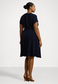 Abito blu navy a maniche corte con vita aderente e gonna a palloncino. Presenta una cerniera sul retro e un tessuto liscio, abbinato a sandali con tacco marroni.