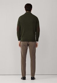 Suéter de punto verde olivo con parches marrones en los codos, combinado con pantalones marrones claros. El suéter tiene un dobladillo y un cuello acanalados.