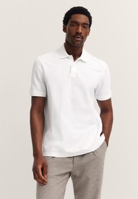 Mann mit kurzen lockigen Haaren, der ein weißes kurzärmeliges Poloshirt und hellgraue Hosen trägt, steht mit einer Hand in der Tasche.