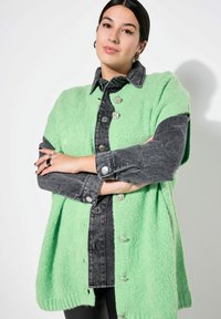 Grön stickad cardigan över en mörk denimskjorta, med silverknappar och en avslappnad passform. Slät textur, avslappnad stil, korta ärmar.