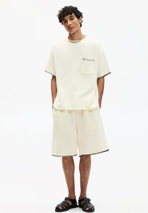 Cremefarvet oversized t-shirt med lomme og kontrastsyning i sort, parret med matchende shorts og sorte sandaler.