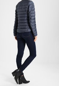 Donna che indossa un giubbotto trapuntato navy, jeans skinny blu scuro e stivaletti neri con tacco a blocco, in piedi contro uno sfondo neutro.