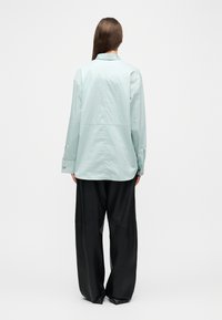 Mujer con cabello largo y castaño, lleva una camisa azul clara de botones y pantalones anchos negros, de pie contra un fondo claro y liso, mirando hacia atrás.