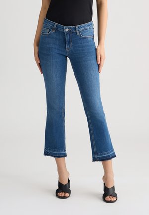 Jeans blu a zampa d'elefante in denim con orlo sfilacciato, caratterizzati da una vita aderente e delicati dettagli di cucitura. Abbinati a tacchi aperti neri.
