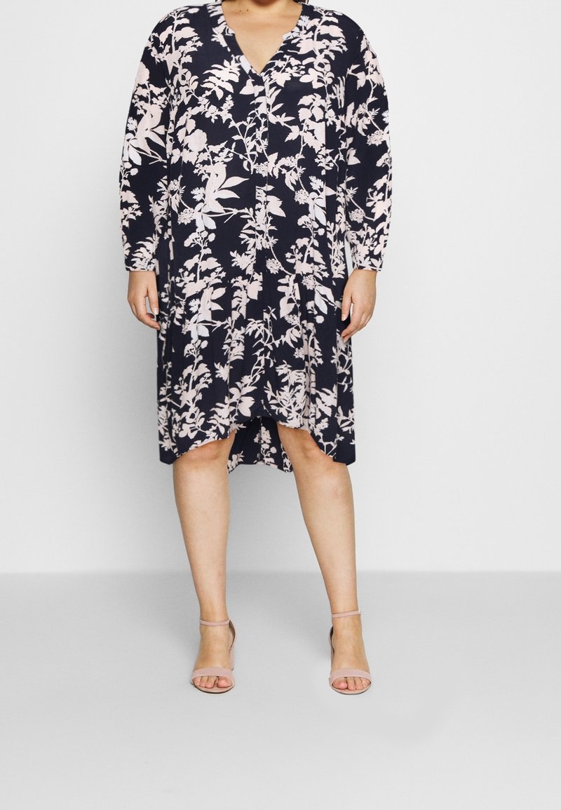 Robe fleurie bleu marine avec des accents roses. Manches longues, décolleté en V et ourlet asymétrique. Assortie à des sandales à lanières rose pâle.