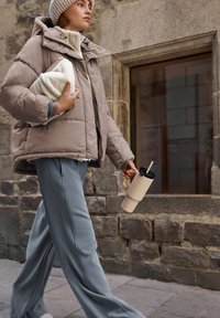 Giacca a vento imbottita color tasso con cappuccio, abbinata a pantaloni grigi a gamba larga. Tiene in mano una tazza riutilizzabile beige e un oggetto bianco con trama.