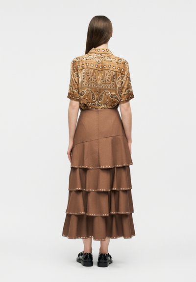 Γυναίκα με μακριά καστανά μαλλιά, φορούσα ένα κοντομάνικο πουκάμισο με σχέδιο και μια στρωματοποιημένη μπεζ φούστα μέσης με διακοσμητικές λεπτομέρειες στα τελειώματα, στραμμένη με την πλάτη σε λευκό φόντο.