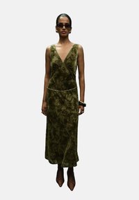Vestido de terciopelo verde oliva con escote en V, cintura ajustada y abertura lateral. Acentuado con un fino cinturón dorado y combinado con tacones puntiagudos.