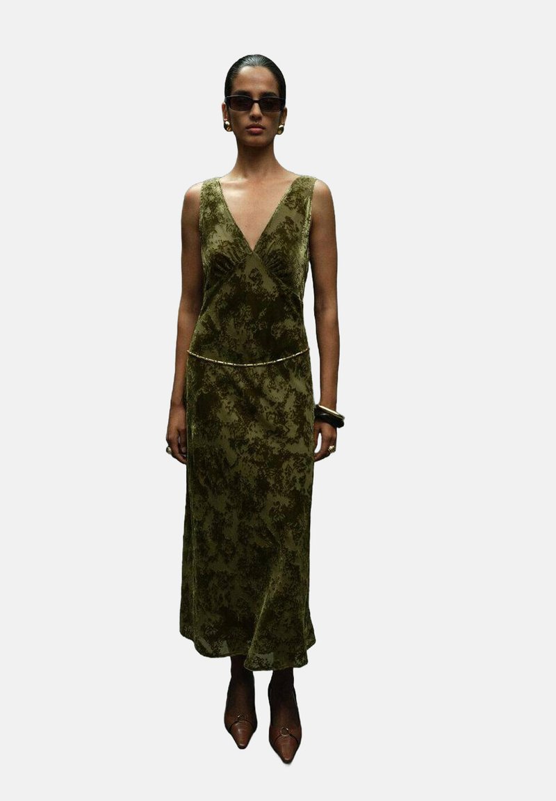 Vestido de terciopelo verde oliva con escote en V, cintura ajustada y abertura lateral. Acentuado con un fino cinturón dorado y combinado con tacones puntiagudos.