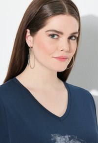 Námořnická modrá v-neck tričko vyrobené z měkké tkaniny, s jemným vzorem u lemování. Modelka má dlouhé vlasy a velké stříbrné kroužkové náušnice.
