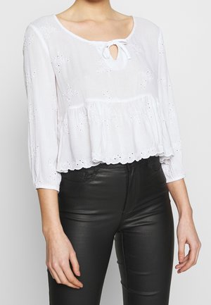 Femme portant un chemisier blanc à manches longues avec broderie ajourée et nœud au col, associé à un pantalon noir ajusté.