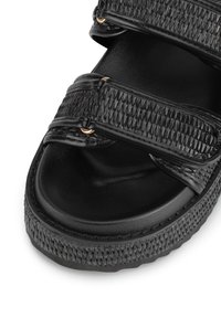Sandal negra con correas sintéticas de textura trenzada, acentos metálicos y suela acolchada. Suela de goma gruesa para mayor durabilidad.