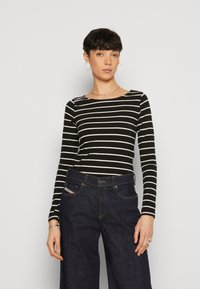 Schwarzes Langarmoberteil mit weißen horizontalen Streifen; runder Ausschnitt. Kombiniert mit dunklen, weit geschnittenen Jeans. Schlichte silberne Schmuckakzente.