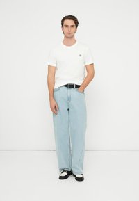 Bílé tričko s krátkým rukávem a logem, světle modré kalhoty s širokými nohavicemi, černý pásek a černobílé boty. Jednoduchý design, pohodlný střih.