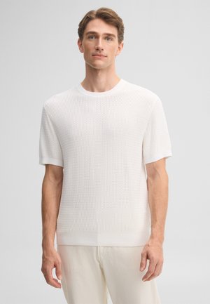 Jeune homme aux cheveux châtains clairs portant une chemise blanche texturée à manches courtes et un pantalon clair, debout devant un fond gris uni.
