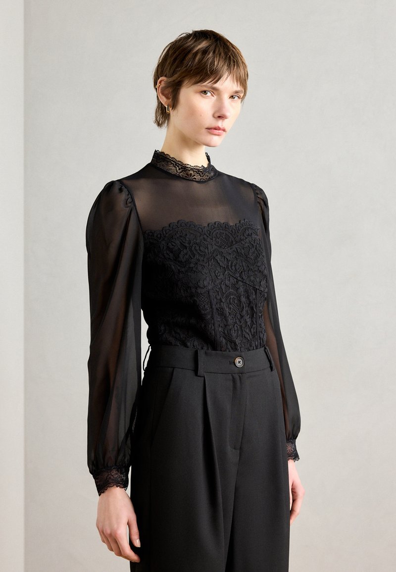 Zwarte blouse met doorzichtige lange mouwen en een hoge kanten kraag, voorzien van ingewikkelde kantdetails op het lijfje, gecombineerd met op maat gemaakte zwarte pantalons.
